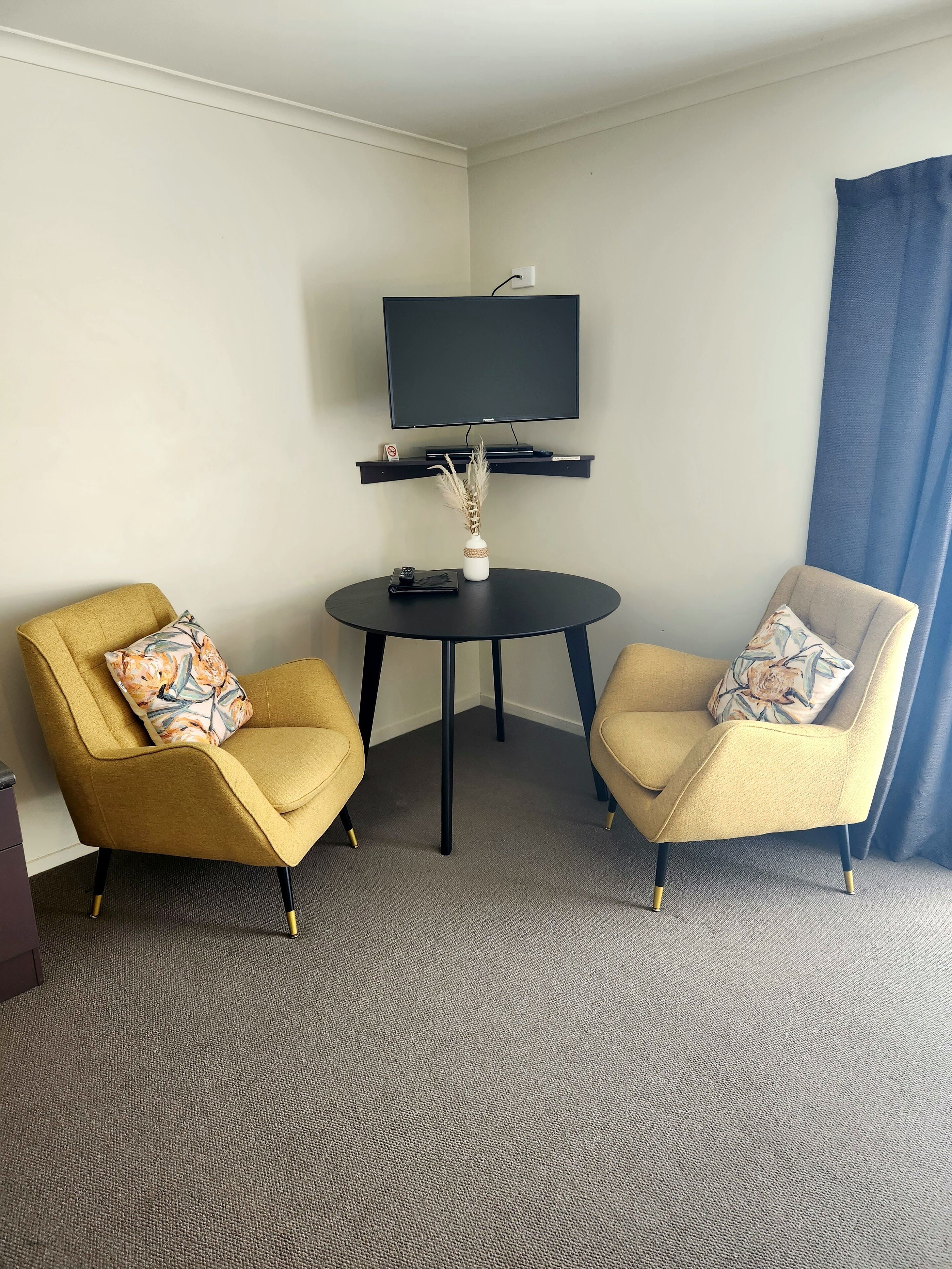 A hotel room with two armchairs, a small round table, a flat-screen TV mounted on the wall, and a vase with dried plants on the table.