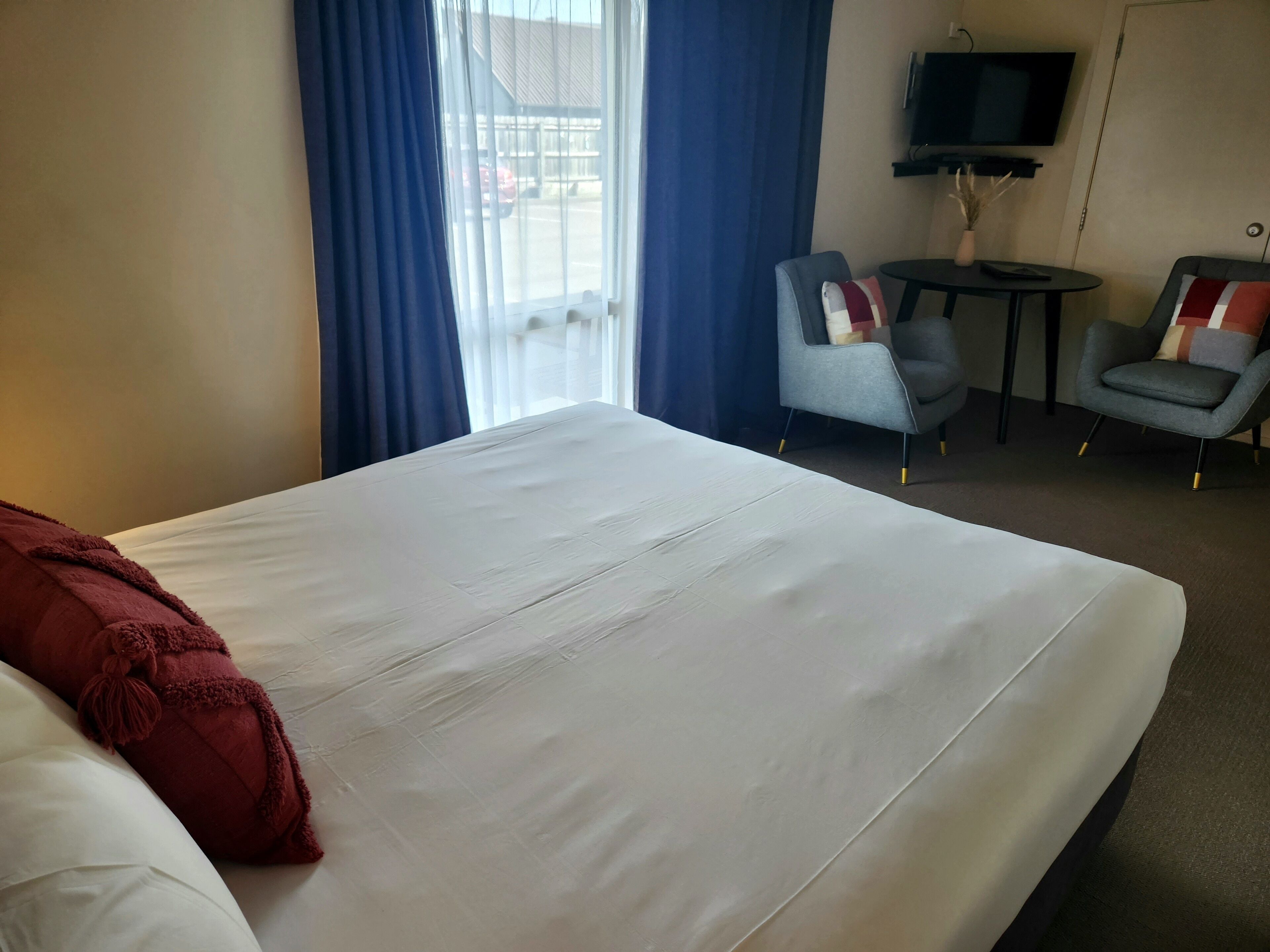 A hotel room with a large bed, two armchairs, a small table, a TV, and a window with curtains.