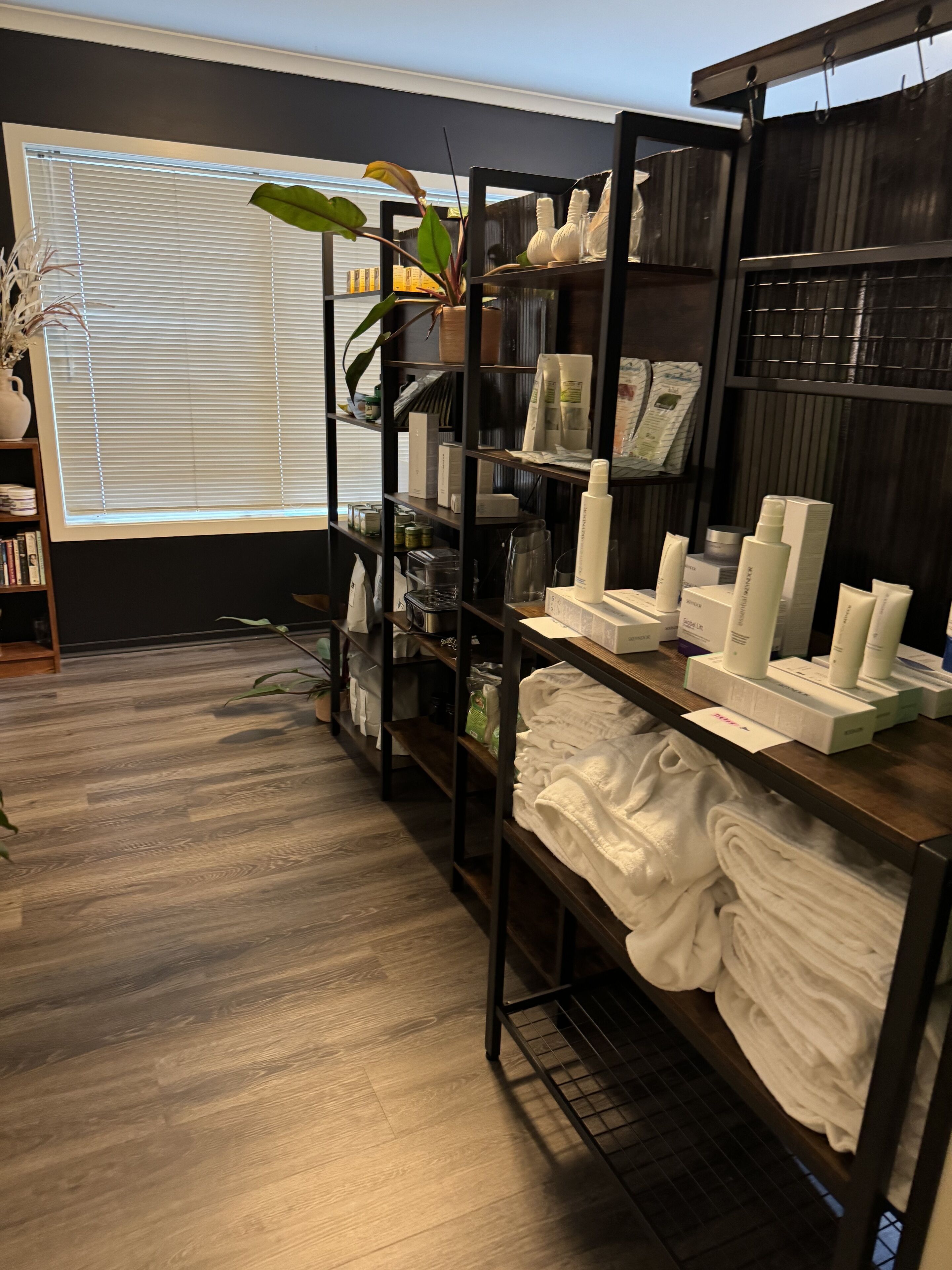 A room with wooden flooring, a window with blinds, a shelf with toiletries, and a plant.