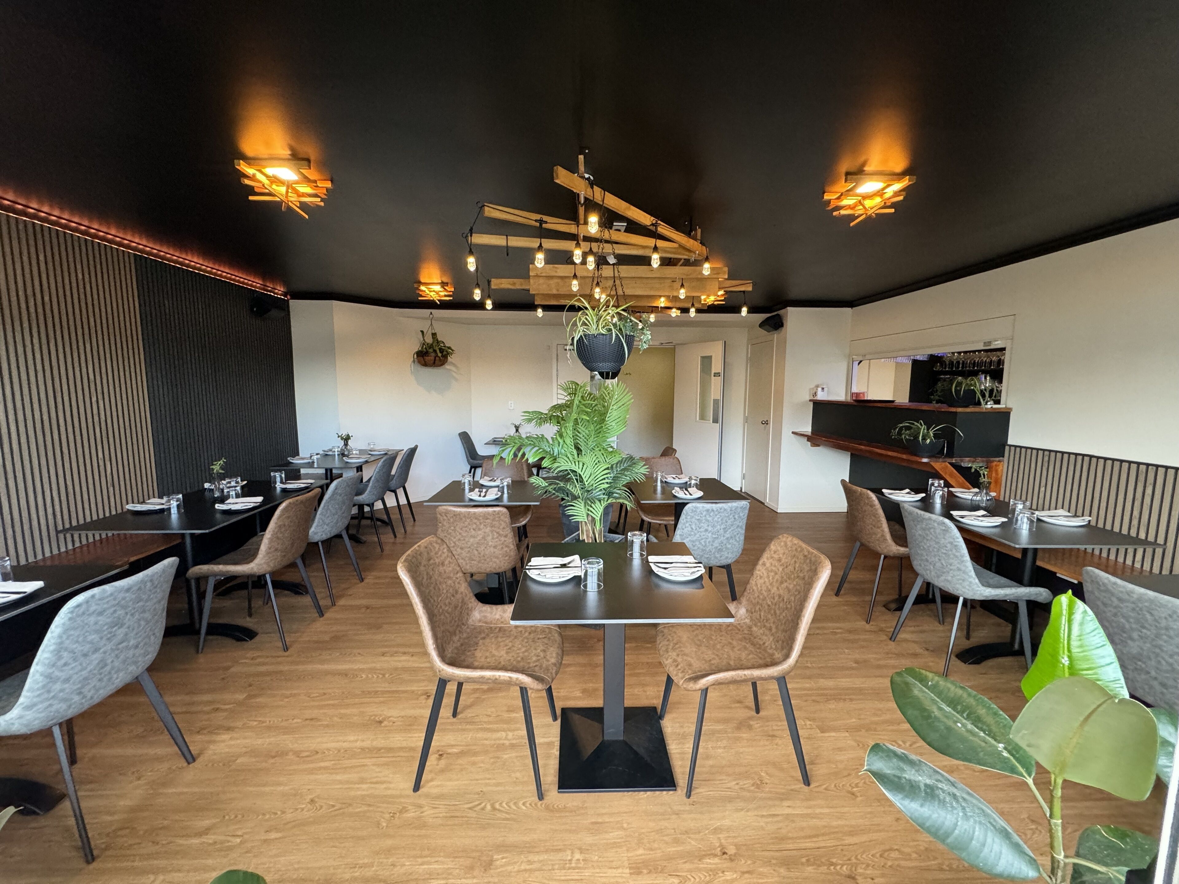 A restaurant interior with wooden tables, grey upholstered chairs, hanging plants, and pendant lighting.