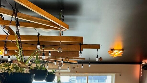 A modern dining area with hanging plants, wooden ceiling beams, and a view of buildings through large windows.