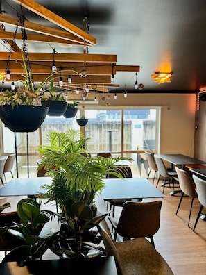 Un comedor moderno con plantas colgantes, vigas de madera en el techo y vistas a edificios a través de amplios ventanales.