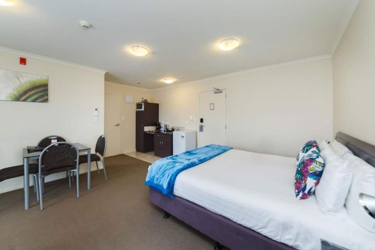 A hotel room with a bed, a desk, a chair, a kitchenette, and a door.