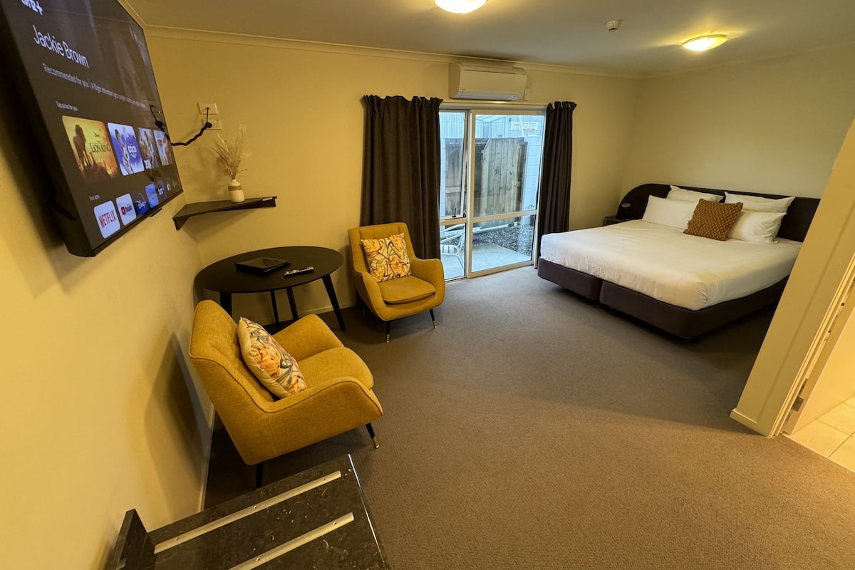 A hotel room with a bed, a sofa, a small table, a TV mounted on the wall, and a sliding glass door leading to a balcony.