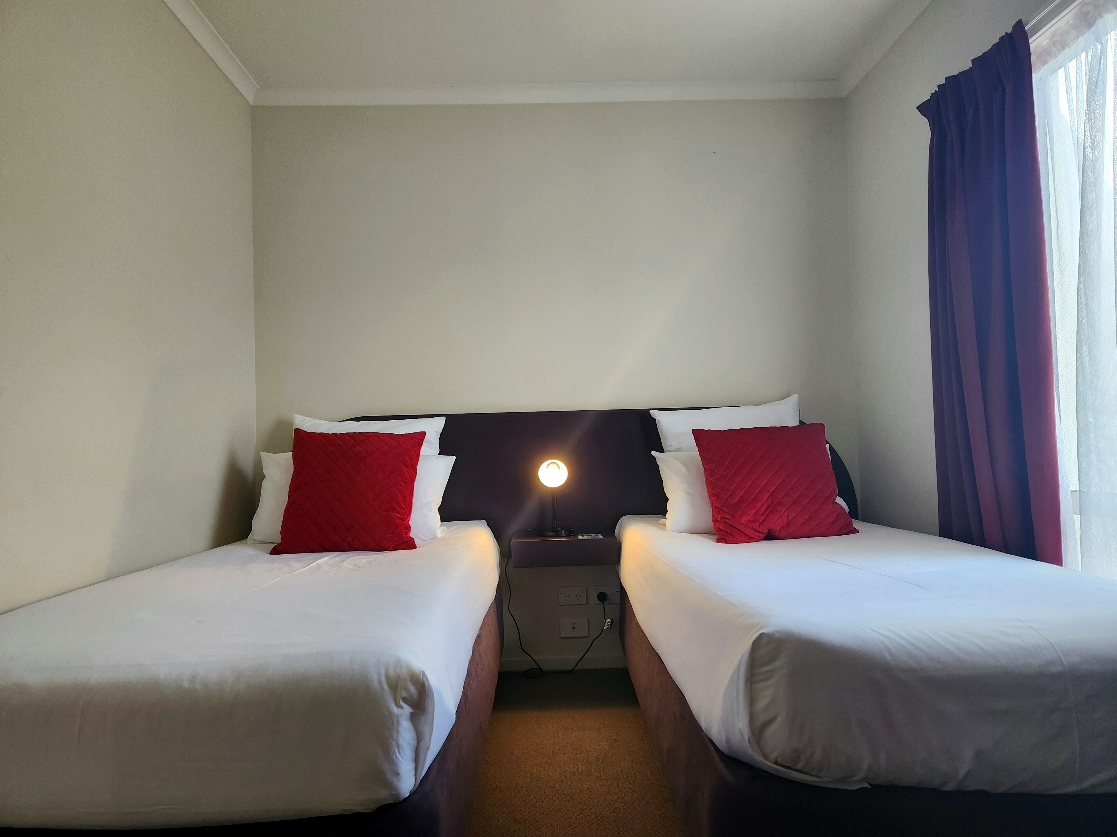 Una camera d'albergo con due letti singoli, entrambi con biancheria da letto bianca e cuscini rossi. Tra i letti si trova un comodino con una lampada. L'ambiente presenta un design semplice e pulito, con una palette di colori neutri.