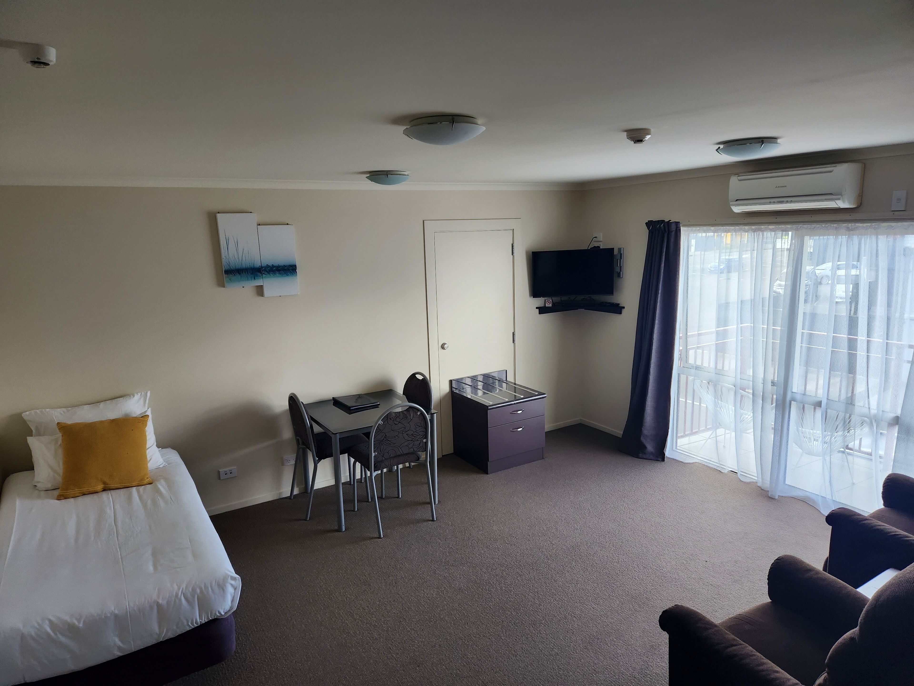 A hotel room with a bed, a desk, a chair, a TV, and an air conditioning unit.