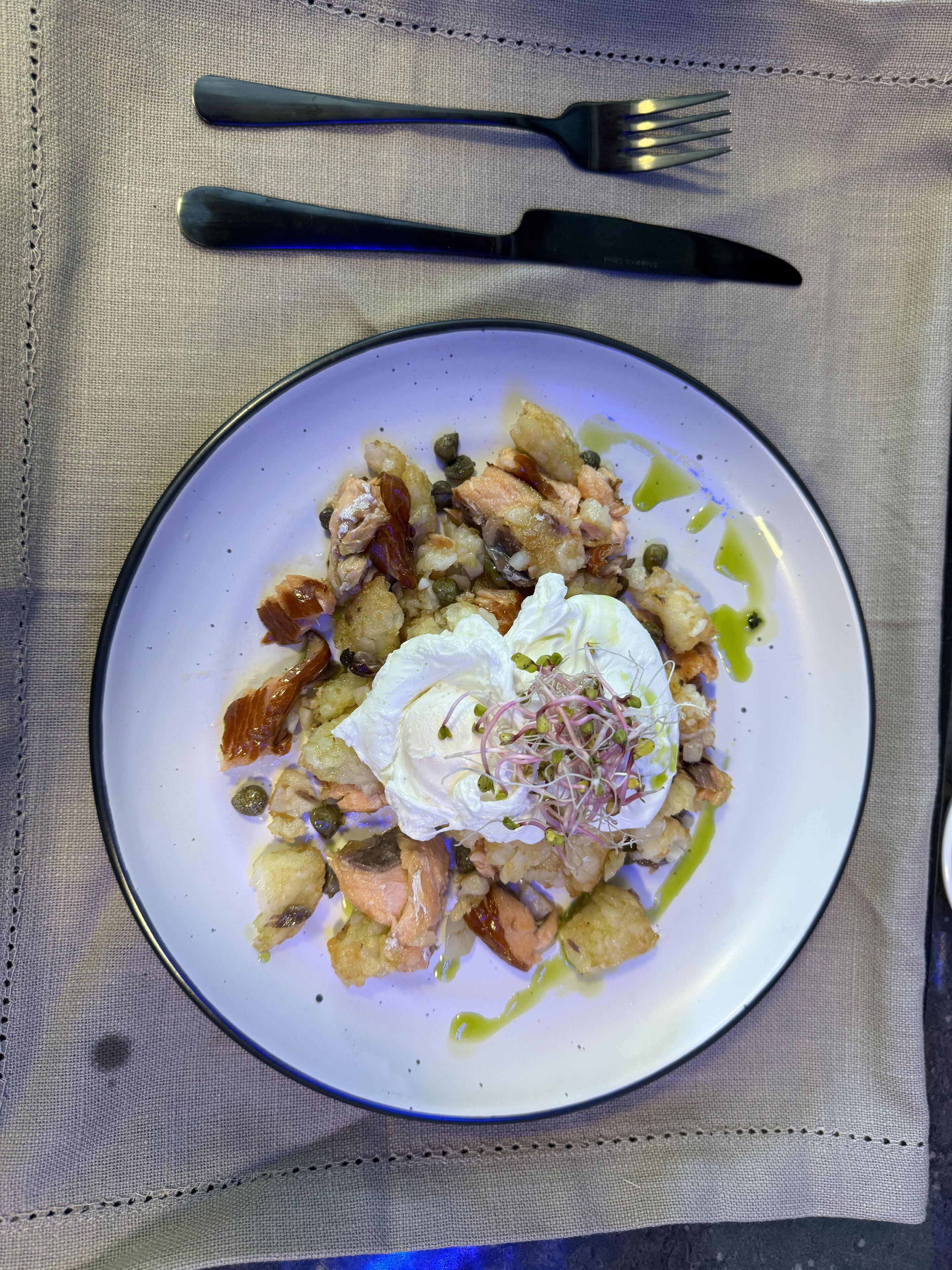 A plate of food with a dollop of cream and sprouts on top, accompanied by a fork and knife on a tablecloth.