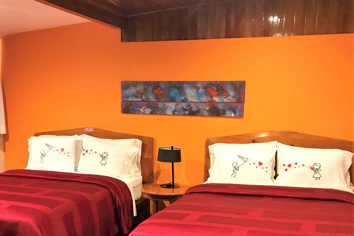Habitación de hotel con dos camas, cada una con una colcha roja y almohadas blancas con diseño de corazón. Hay un cuadro en la pared sobre las camas y una lámpara en la mesita de noche.