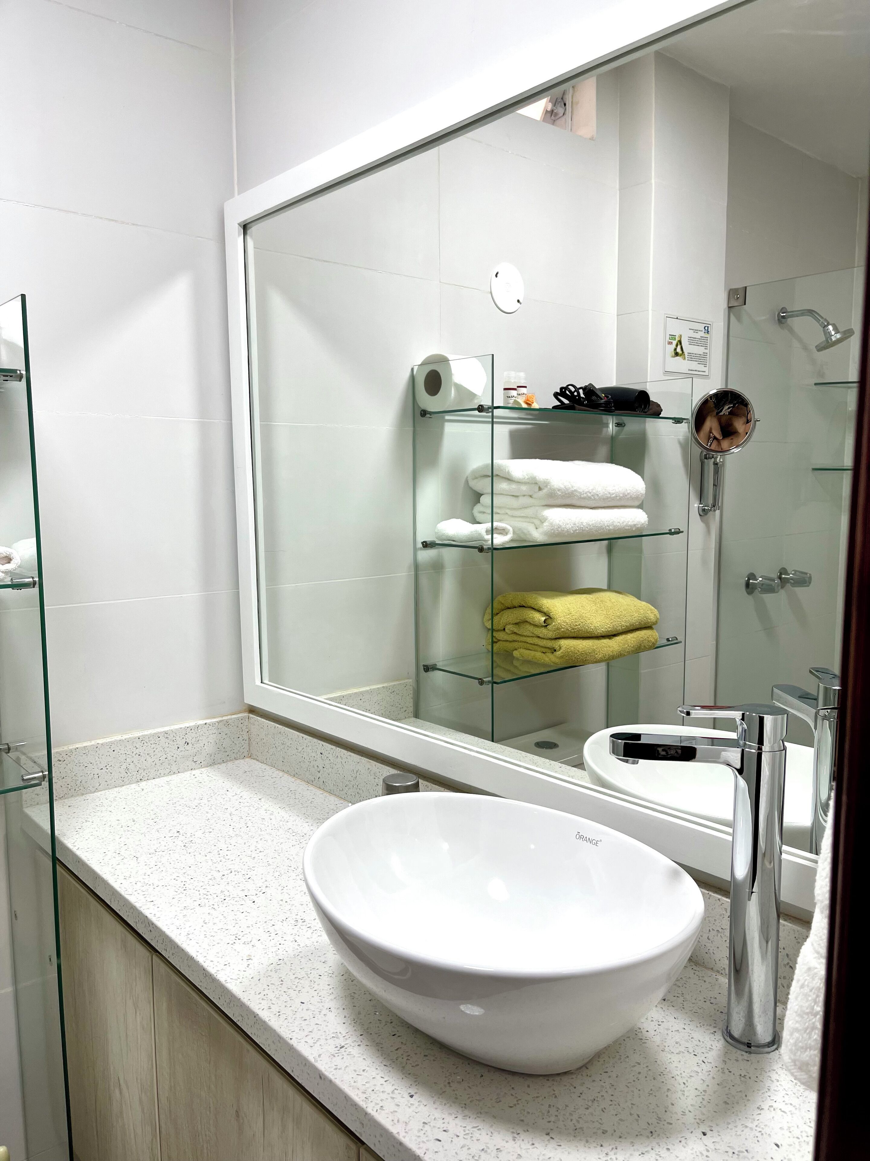 Ein modernes Badezimmer mit einem großen Spiegel, einem weißen Waschbecken und Glastregalen mit Handtüchern.