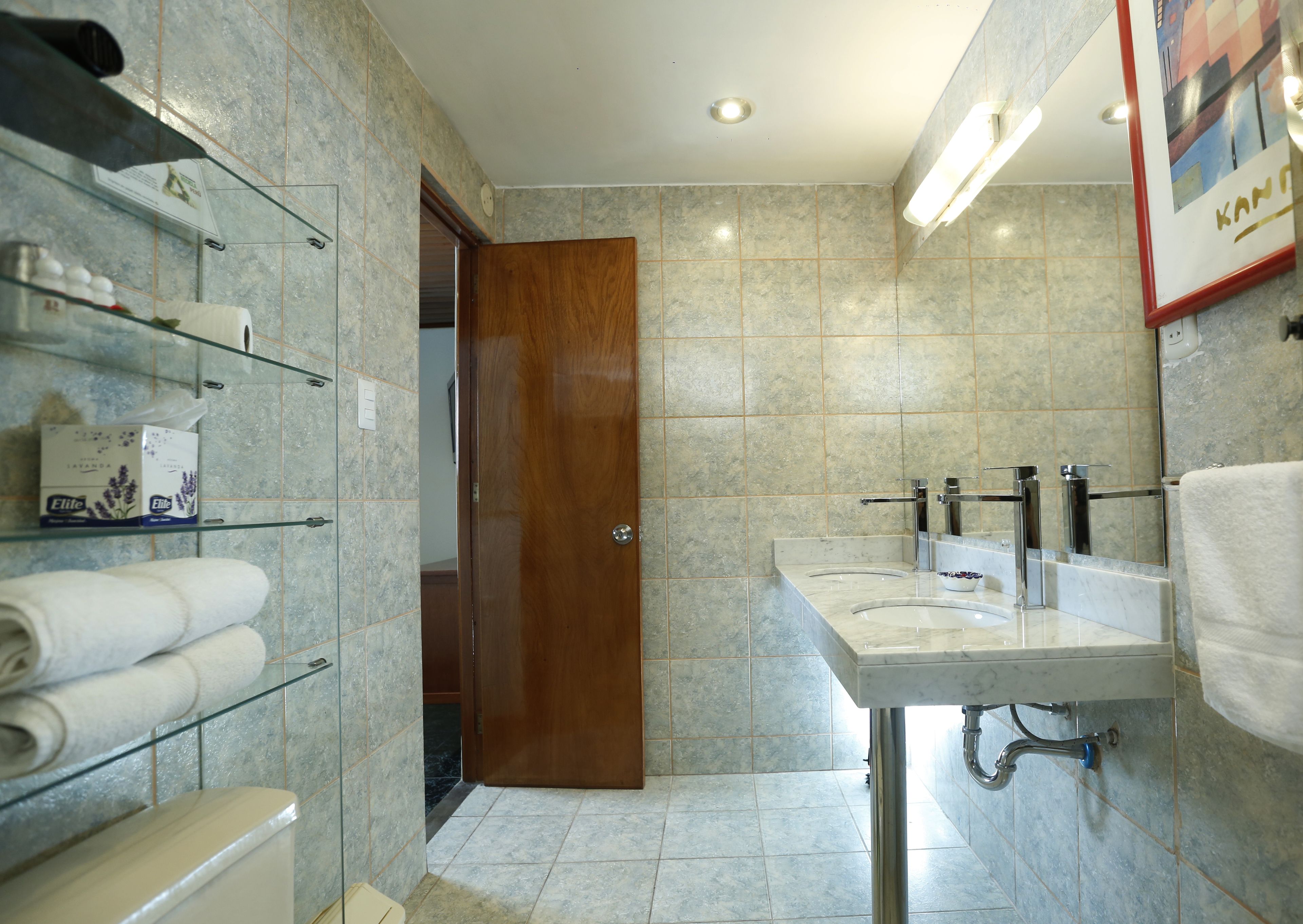 Baño con piso de mármol, puerta de madera, repisa de vidrio con artículos de aseo y lavamanos con espejo.