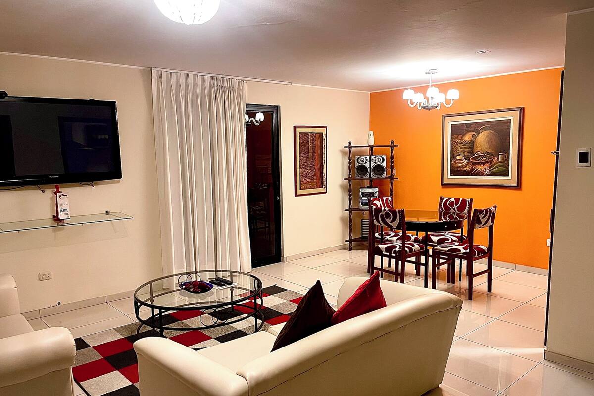 Una sala de estar con sofá, mesa de centro, televisor, comedor con sillas y mesa, y una araña.