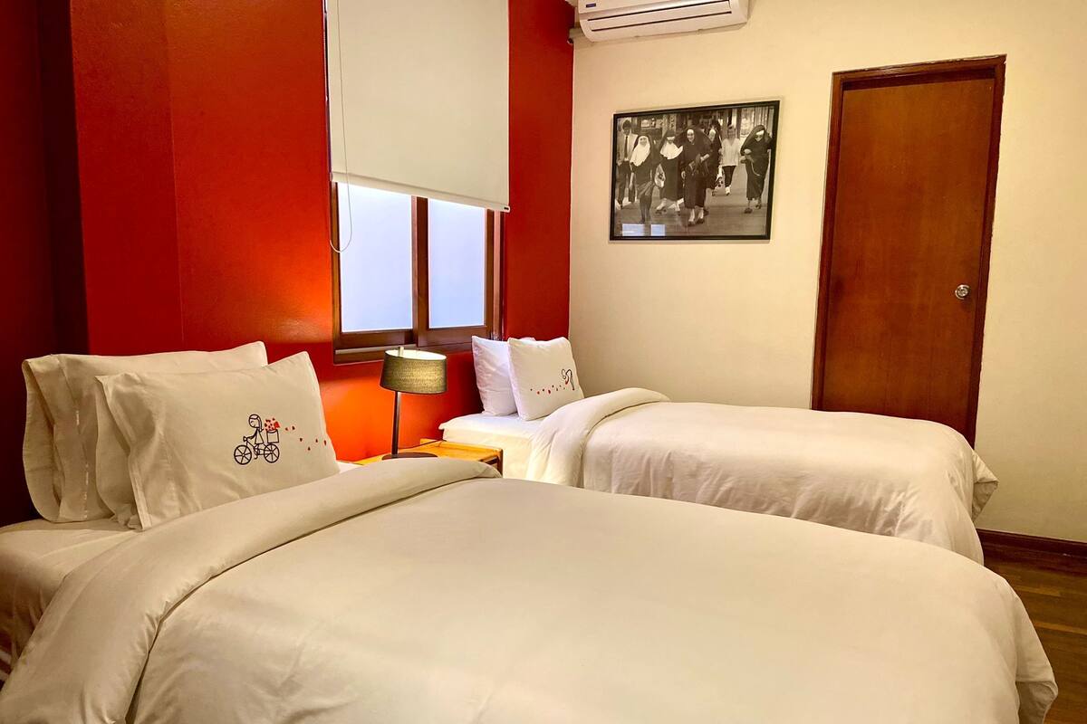 Habitación de hotel con dos camas, una ventana con persiana blanca, una lámpara en una mesita de noche y un cuadro en la pared.