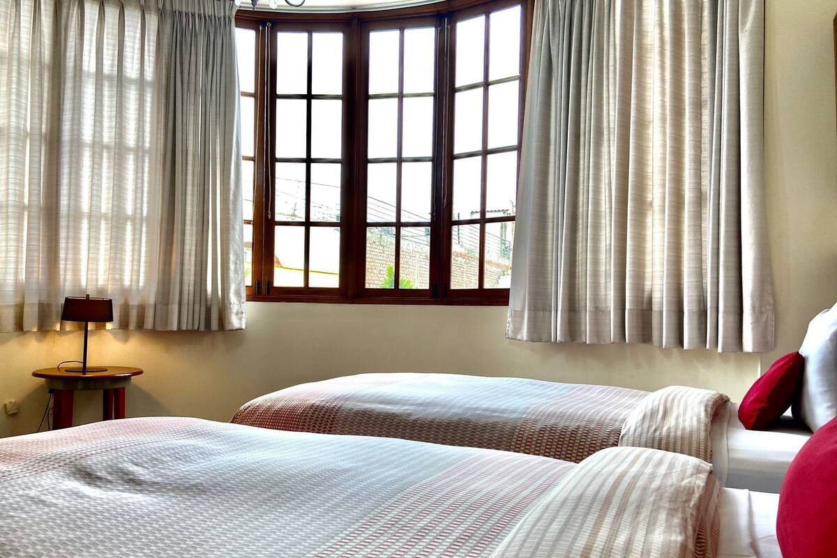 Habitación de hotel con dos camas, una ventana con cortinas, una lámpara en una mesita de noche y vistas al exterior.