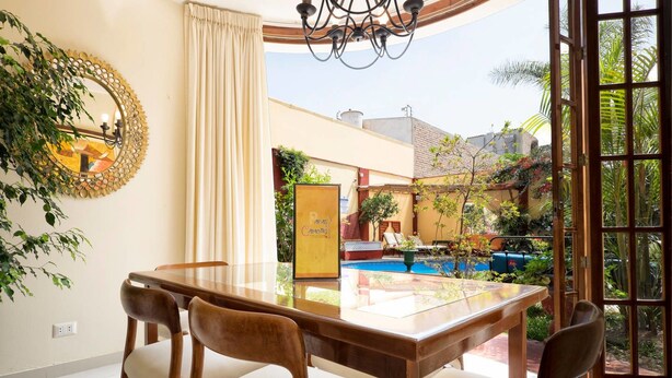 Un comedor con mesa de vidrio, cuatro sillas, un espejo decorativo y vistas a una piscina exterior y jardín.