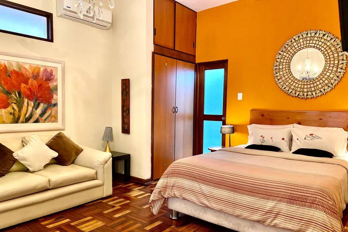 Habitación de hotel con suelo de madera tipo espiga, cama con colcha estampada, sofá blanco con cojines, un cuadro en la pared y un espejo decorativo sobre la cama.