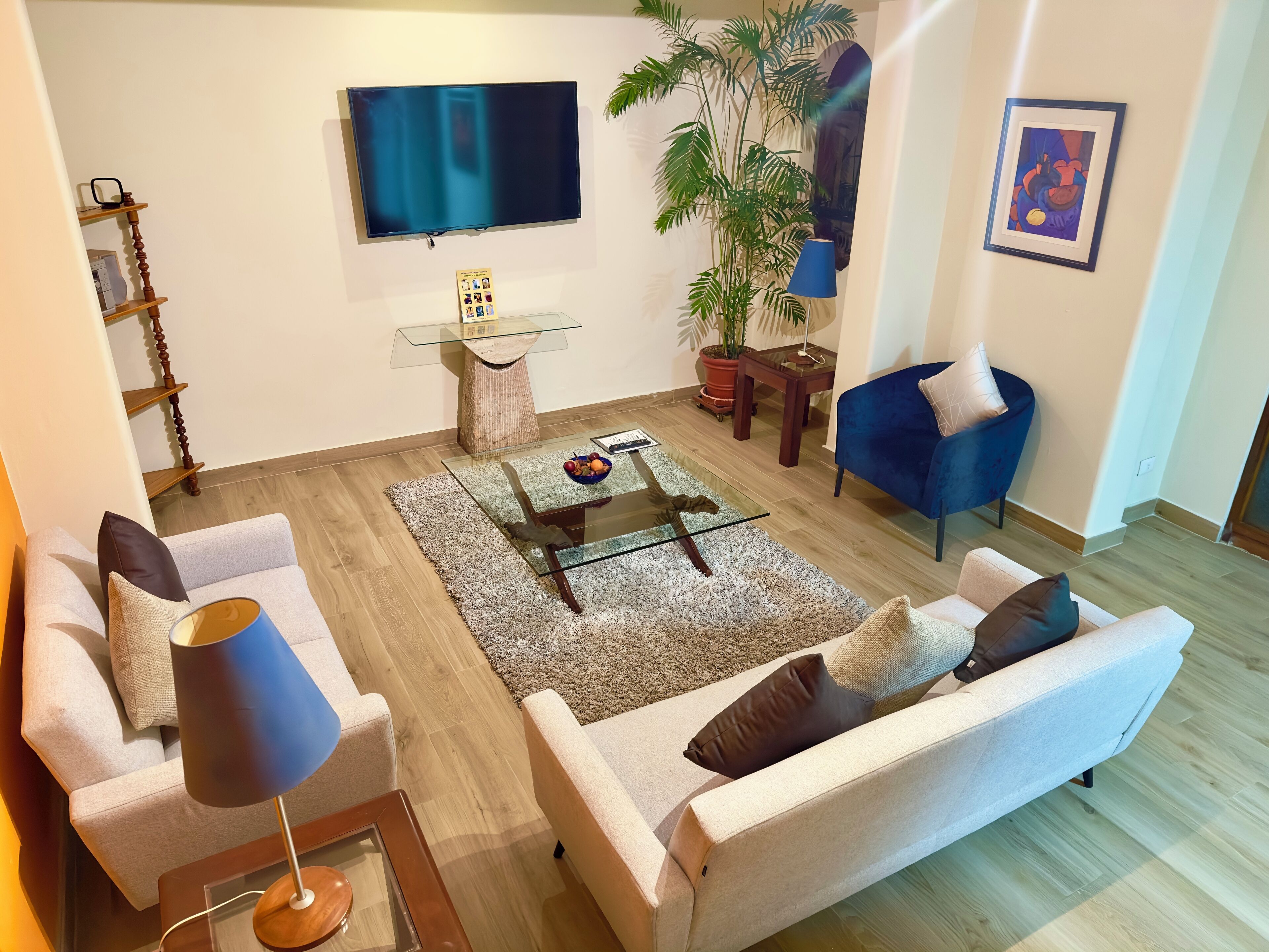 Un saló modern amb televisió de pantalla plana, una taula de cafè de vidre, un butac armchair blau, un sofà beix i una planta gran en test.