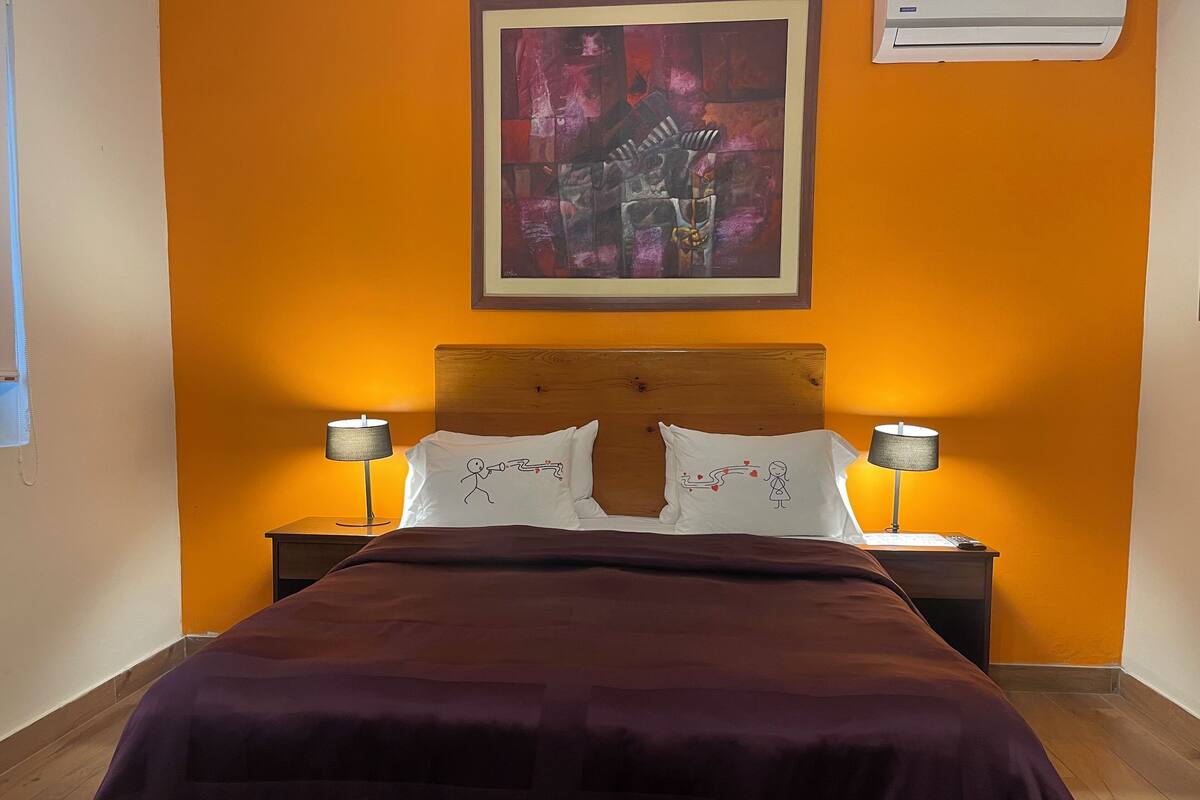 Un cuarto de hotel con cabecera de madera, dos lámparas de noche, una cama con cubrecama morado y un cuadro abstracto enmarcado en la pared.