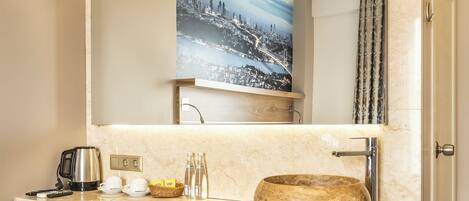 A bathroom with a large mirror, a sink with a stone basin, a coffee maker, and a framed cityscape picture above the mirror.