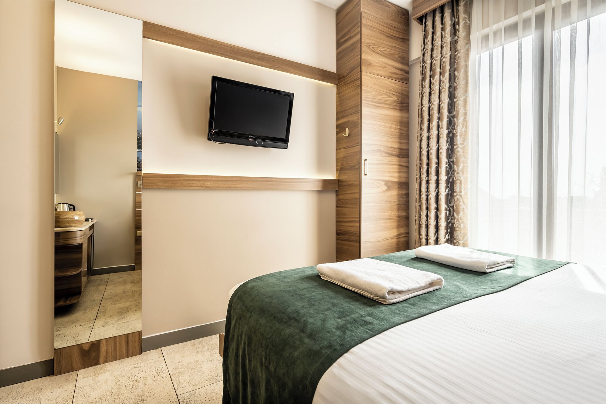 Hotelkamer met een bed, een aan de muur bevestigde televisie, een badkamer met een wastafel en spiegel, en een raam met gordijnen.