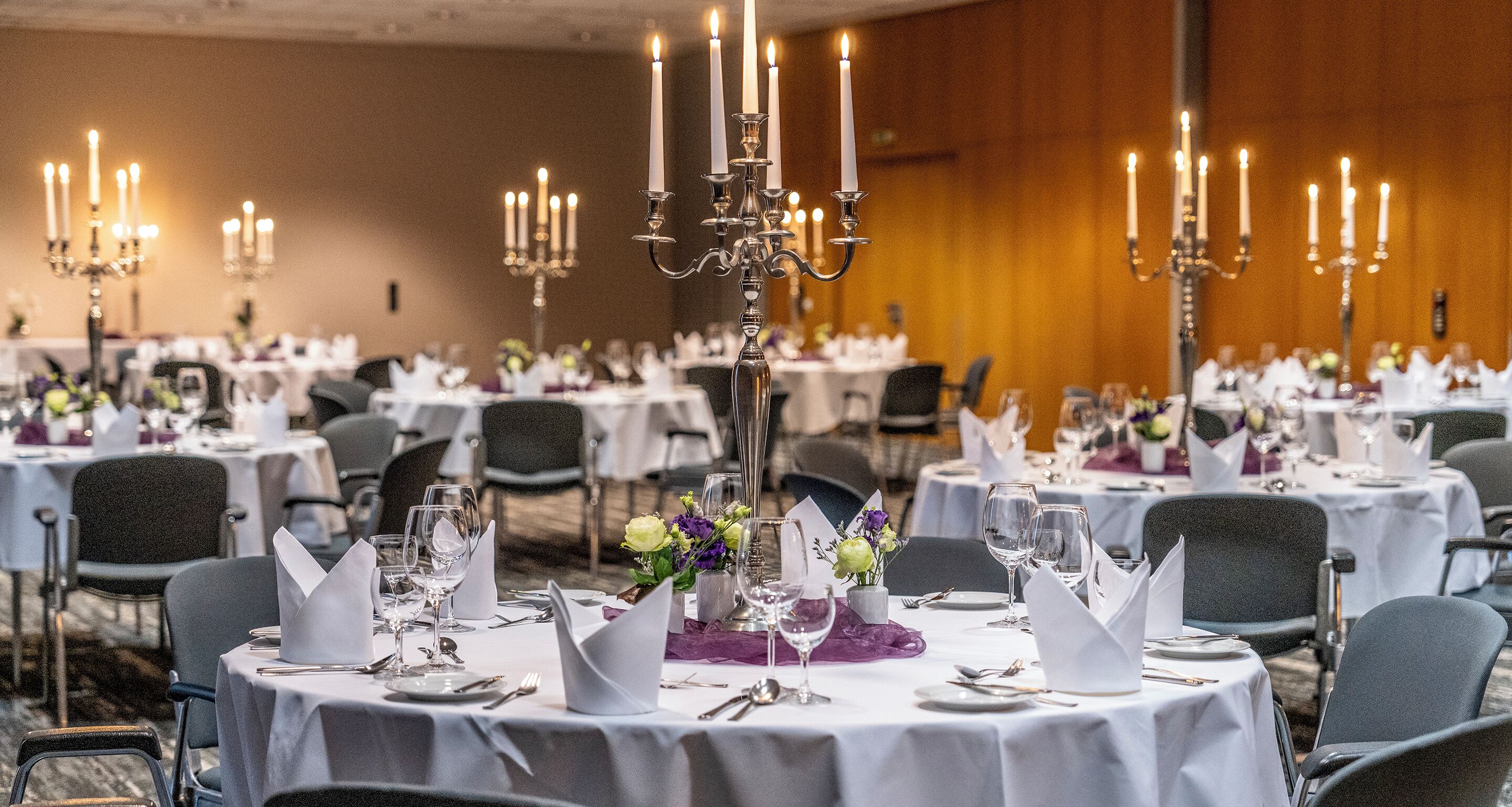 A banquet hall with round tables set for dining, each with white tablecloths, silverware, and candle-lit candelabras.