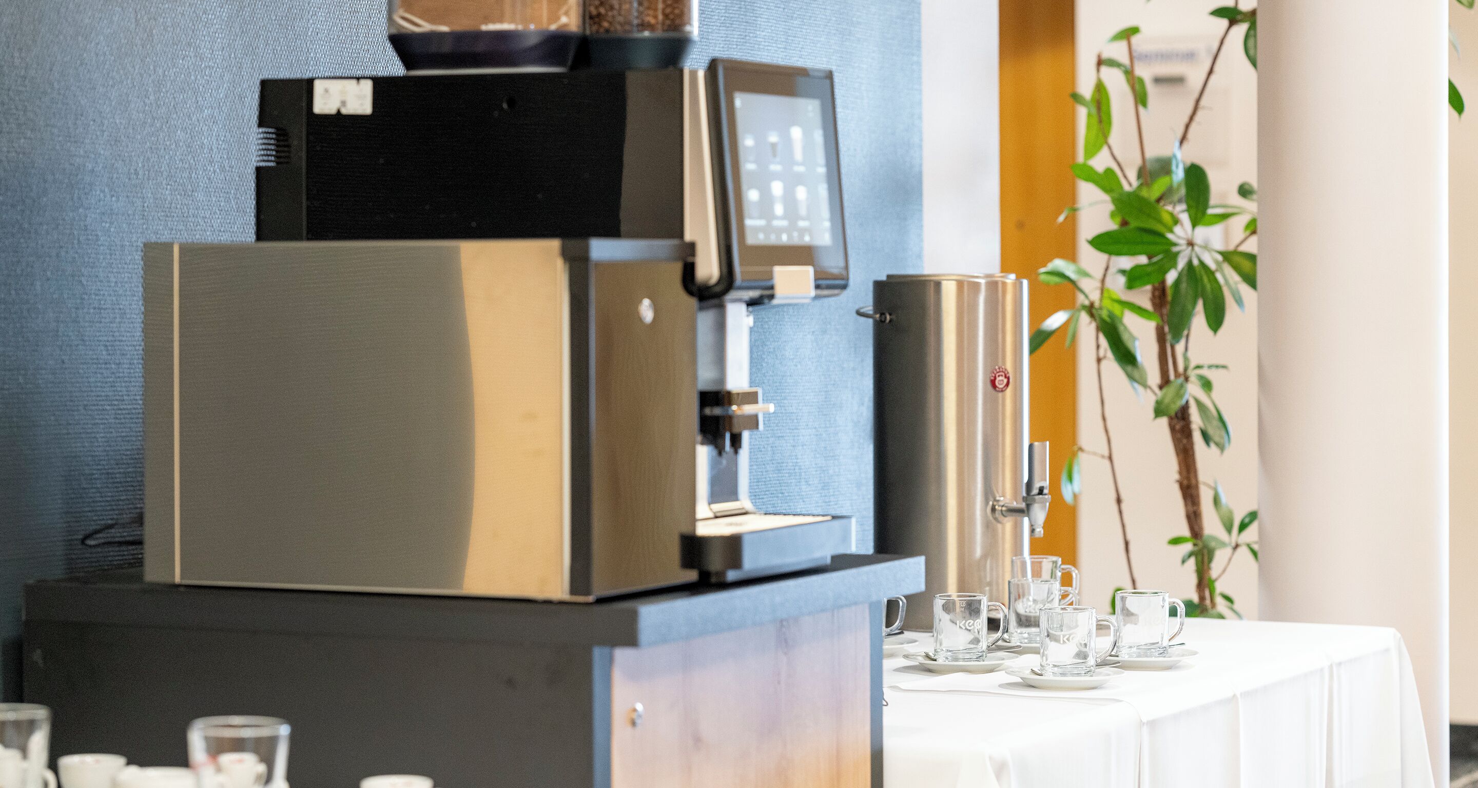 A coffee machine and a water dispenser on a counter, with a table set with clear glasses and saucers.