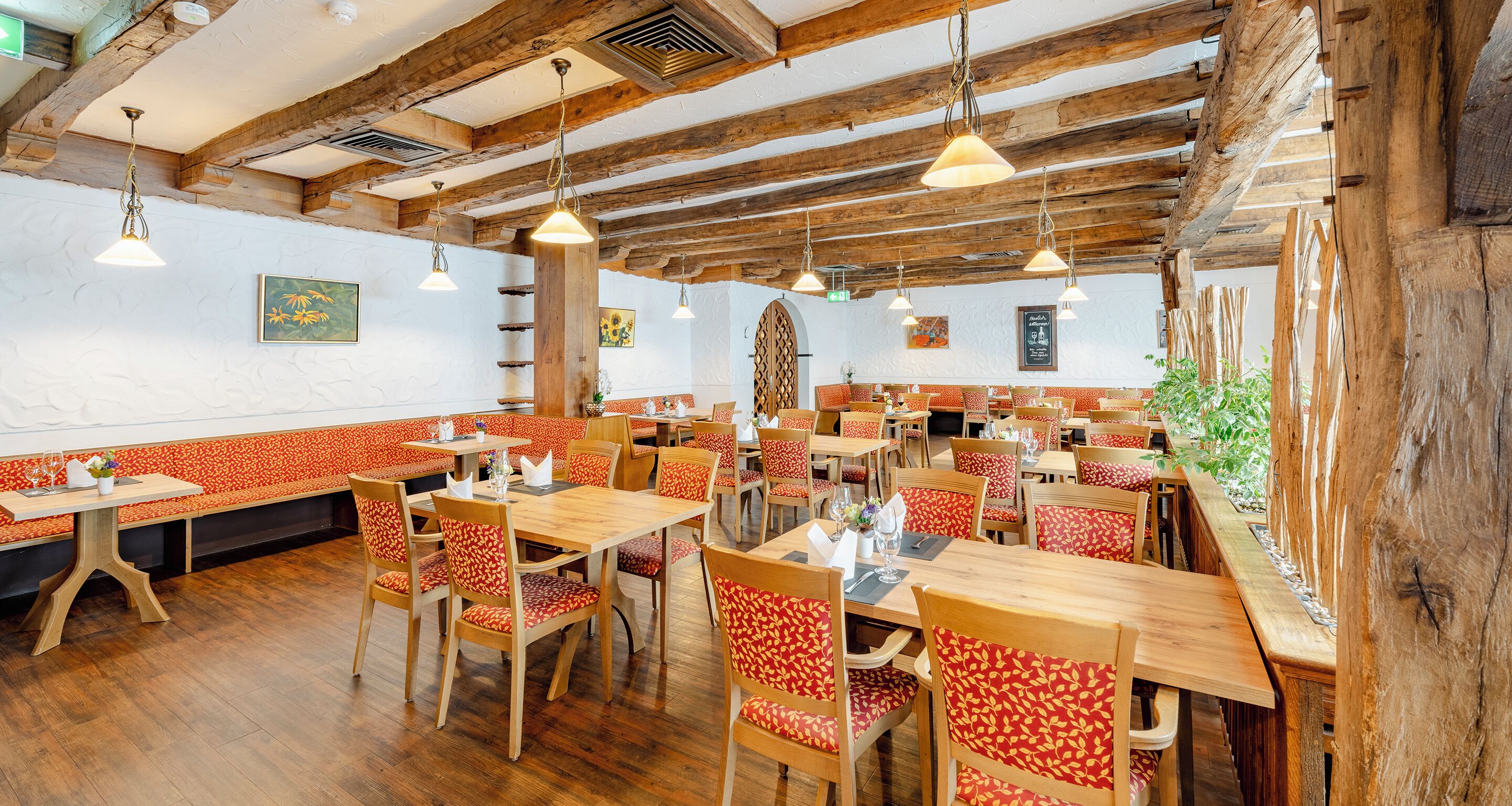 Un restaurant avec des tables et des chaises en bois, un plafond haut avec des poutres apparentes et des luminaires suspendus.