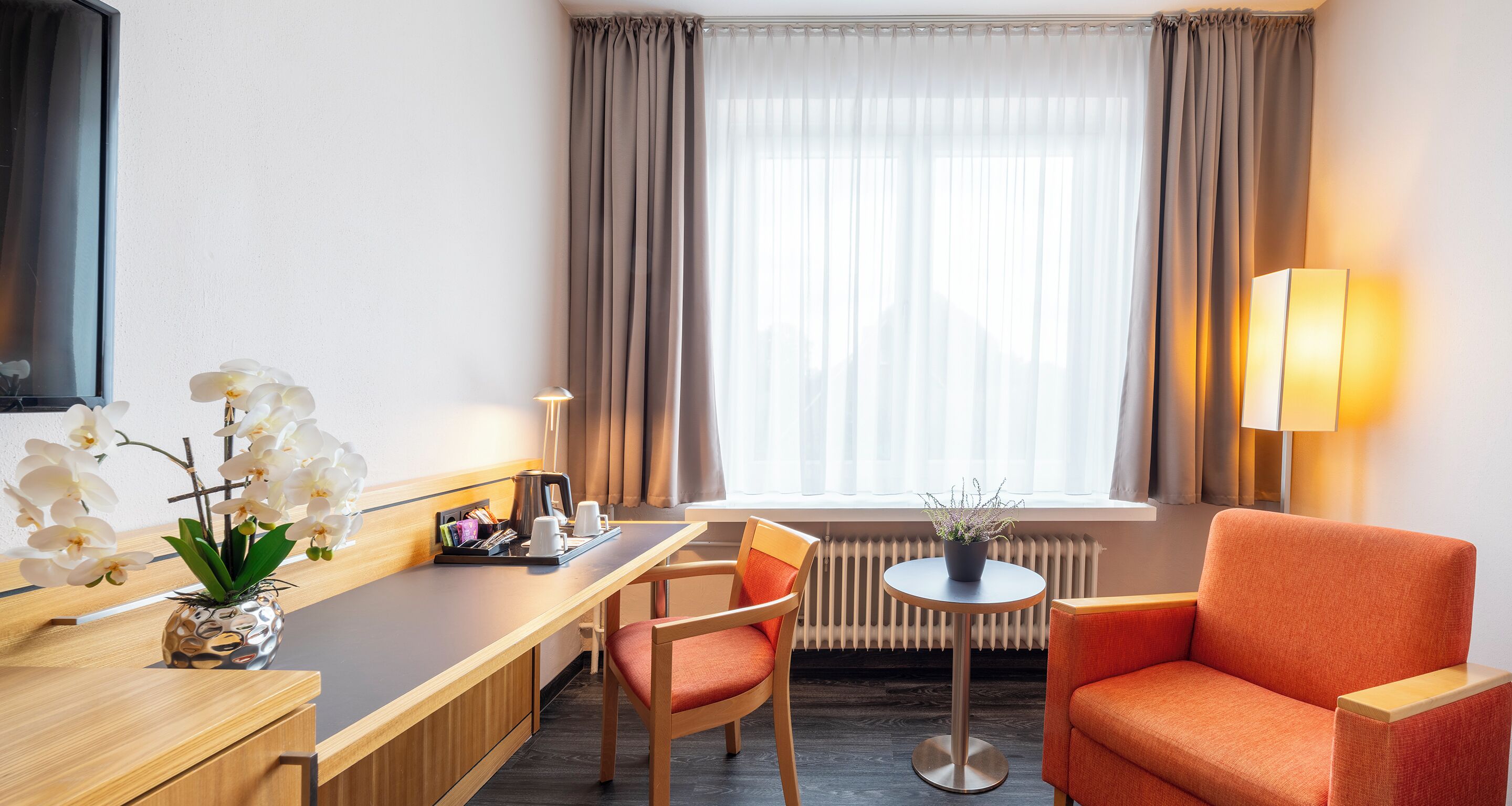 A hotel room with a wooden desk, a chair, a small table with a plant, a lamp, and a window with curtains.