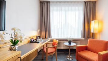 Habitación de hotel con un escritorio de madera, una silla, una mesita con una planta, una lámpara y una ventana con cortinas.