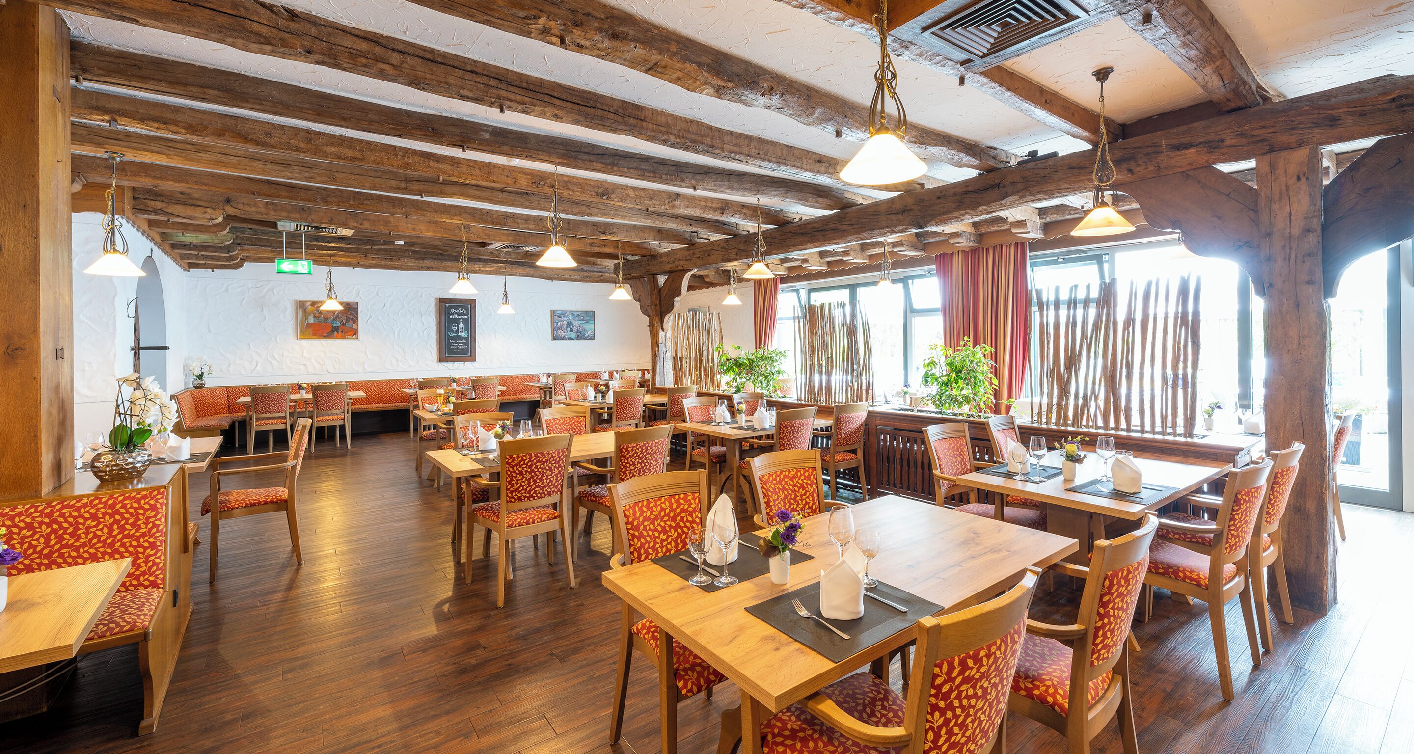 A restaurant with wooden beams, tables with orange patterned chairs, and hanging pendant lights.
