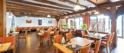 A restaurant with wooden beams, tables with orange patterned chairs, and hanging pendant lights.