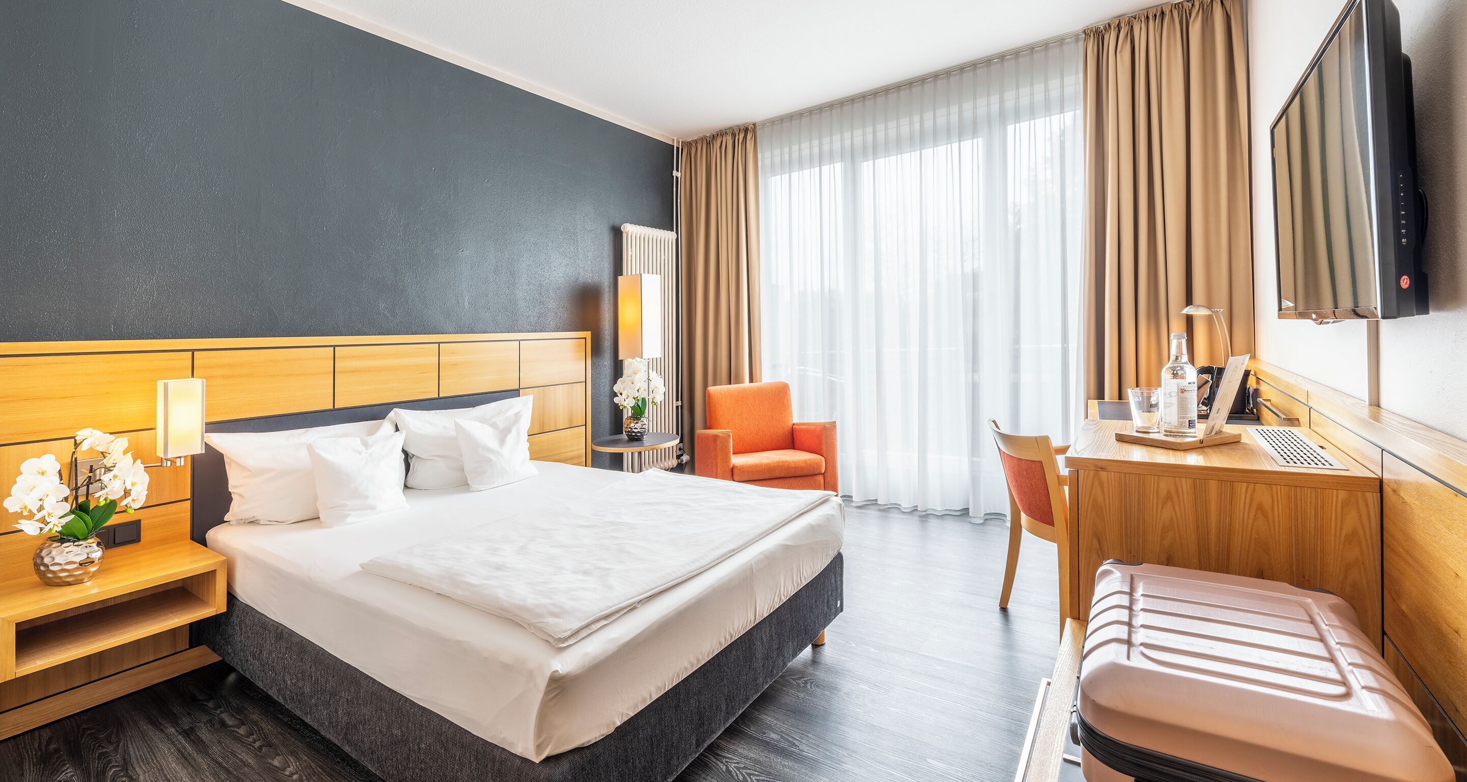 A hotel room with a large bed, a desk with a computer, a chair, and a TV mounted on the wall.