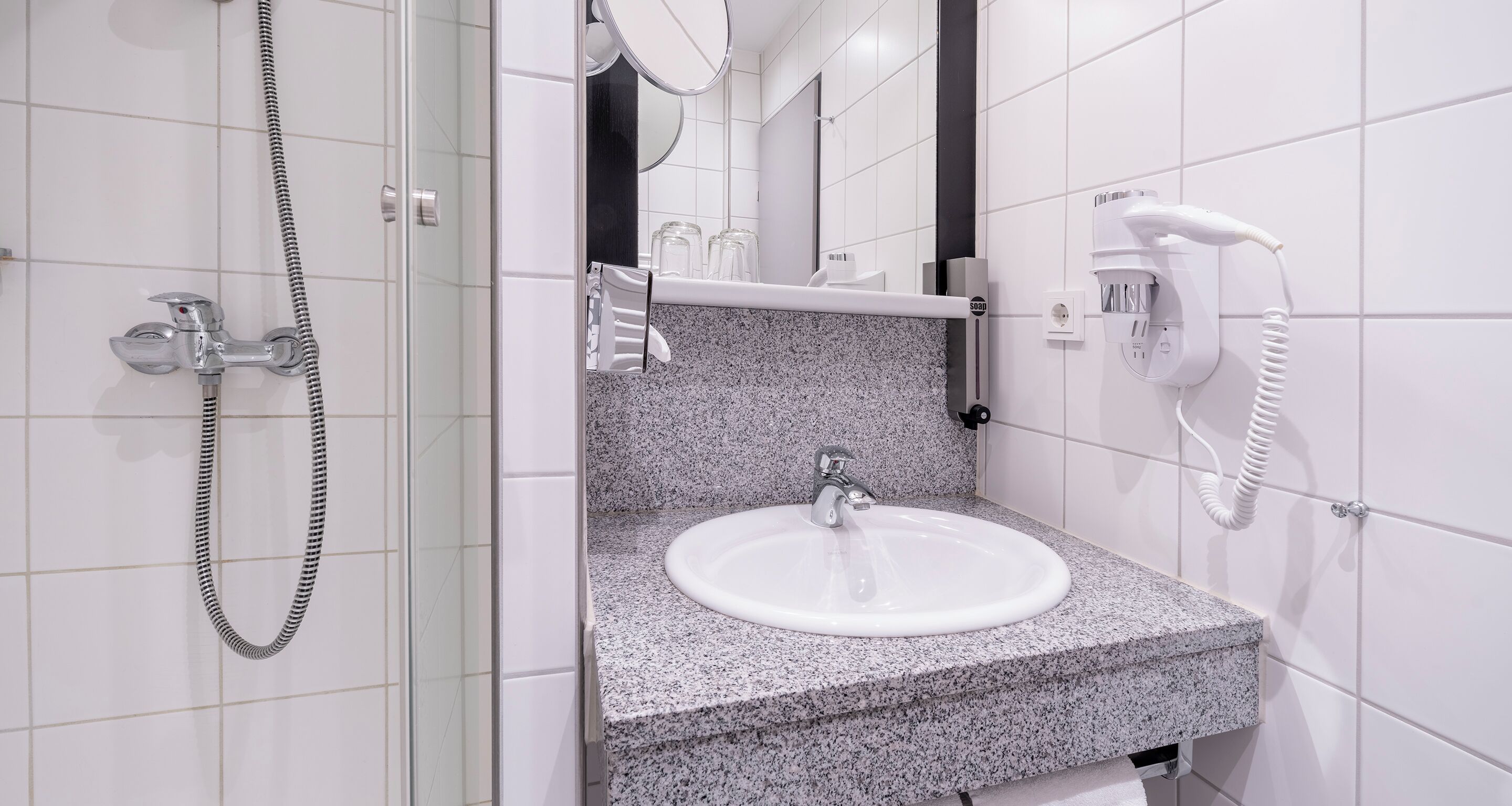 A bathroom with a granite countertop sink, a handheld showerhead, a towel rack, and a hairdryer mounted on the wall.