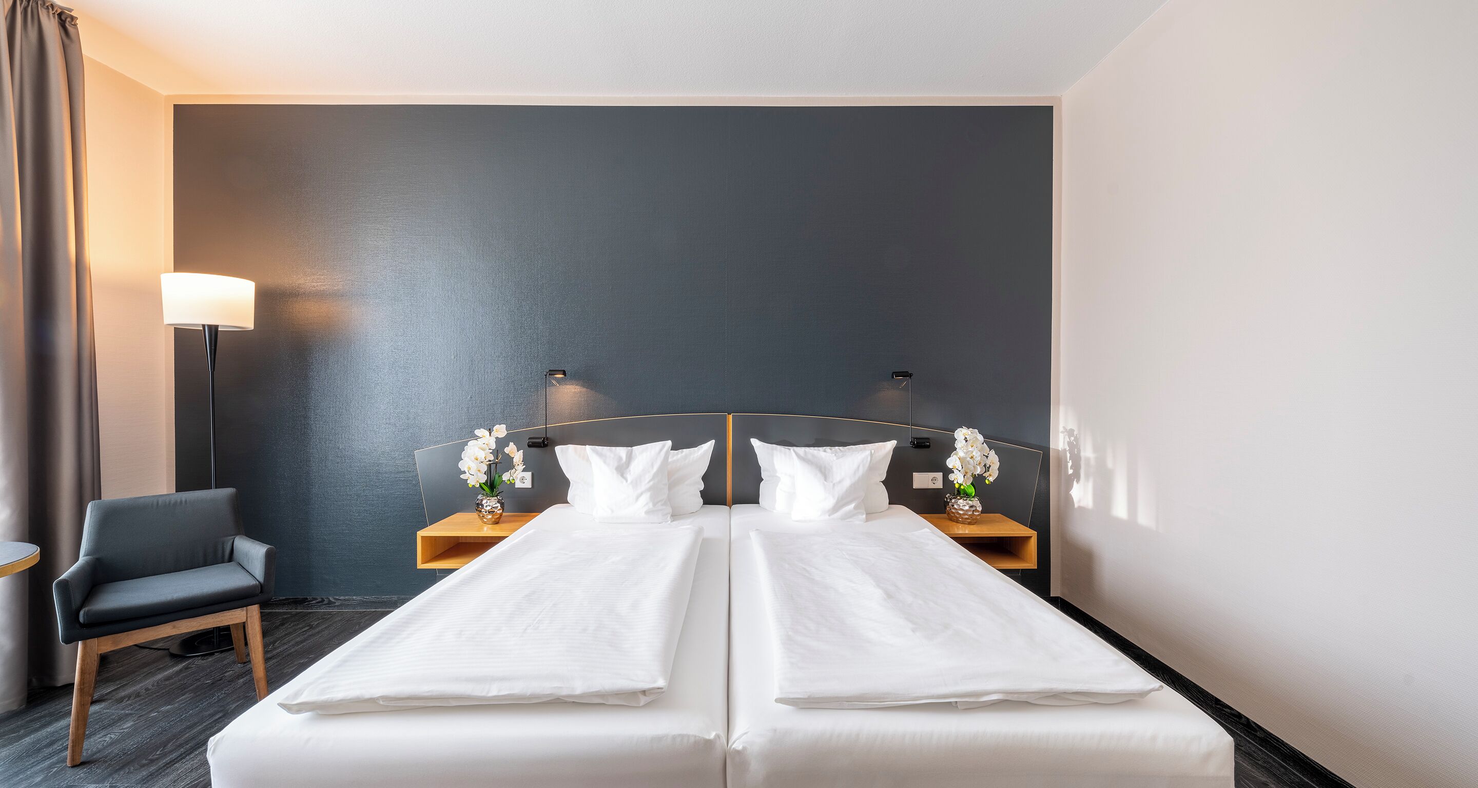 Habitación de hotel con una cama doble, una silla, una mesita de noche con lámpara y un jarrón con flores.