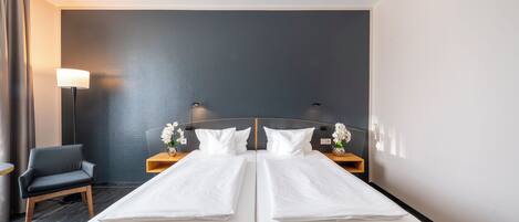 Habitación de hotel con una cama doble, una silla, una mesita de noche con lámpara y un jarrón con flores.