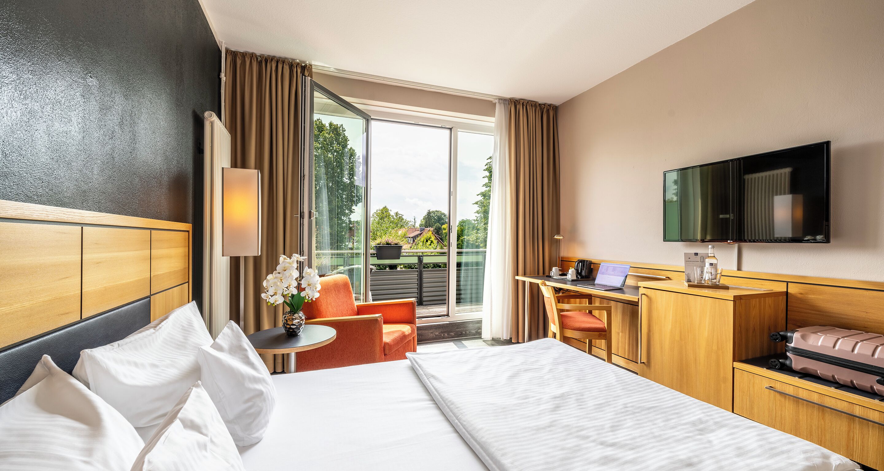 A hotel room with a bed, a desk with a laptop, a television, and a view of greenery outside.