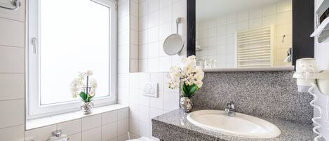 Um banheiro moderno com pia de bancada em granito, vaso sanitário, janela com flores brancas e espelho.