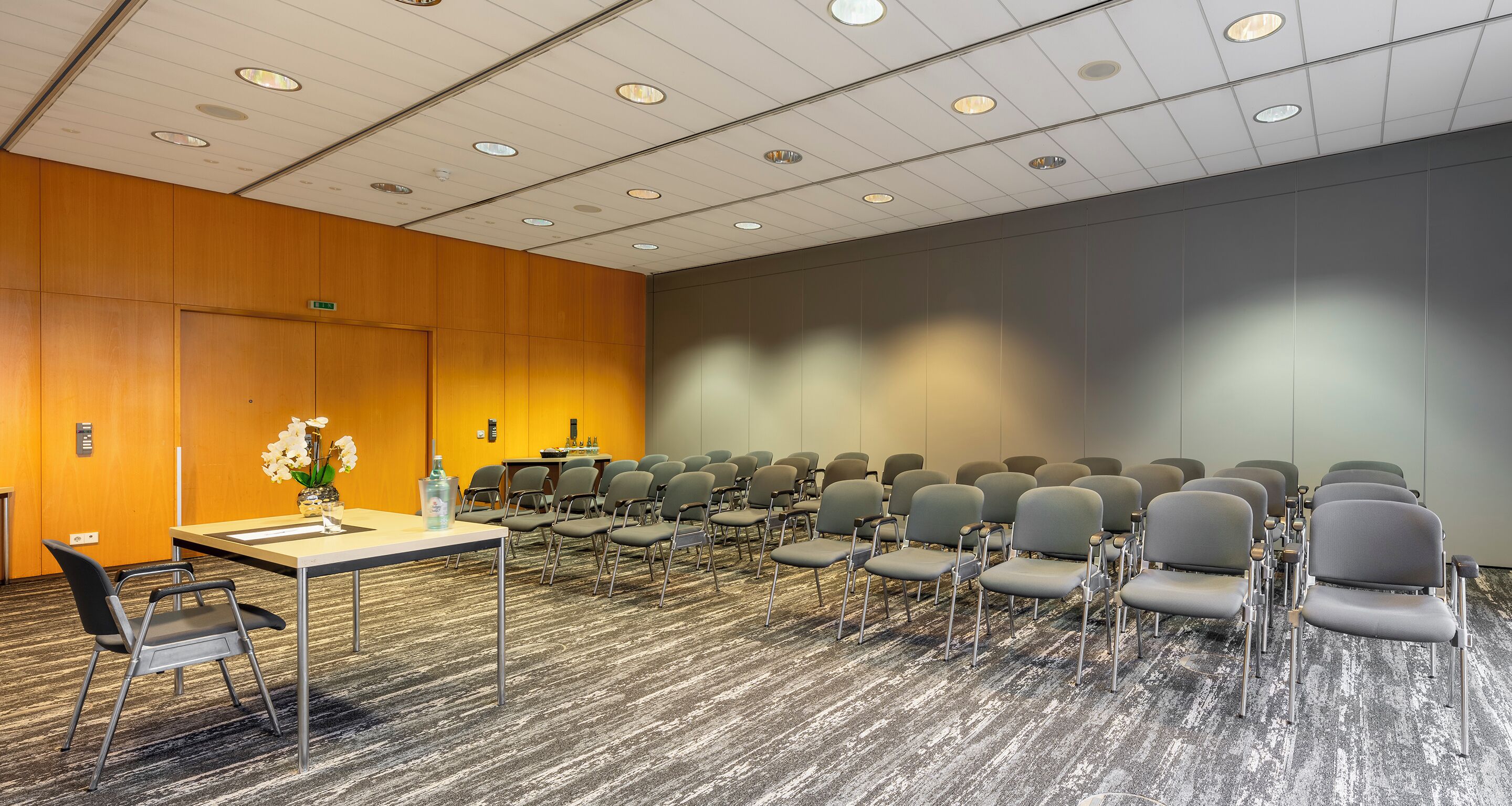 Un salón de conferencias con filas de sillas, una mesa con un florero y una puerta de madera.