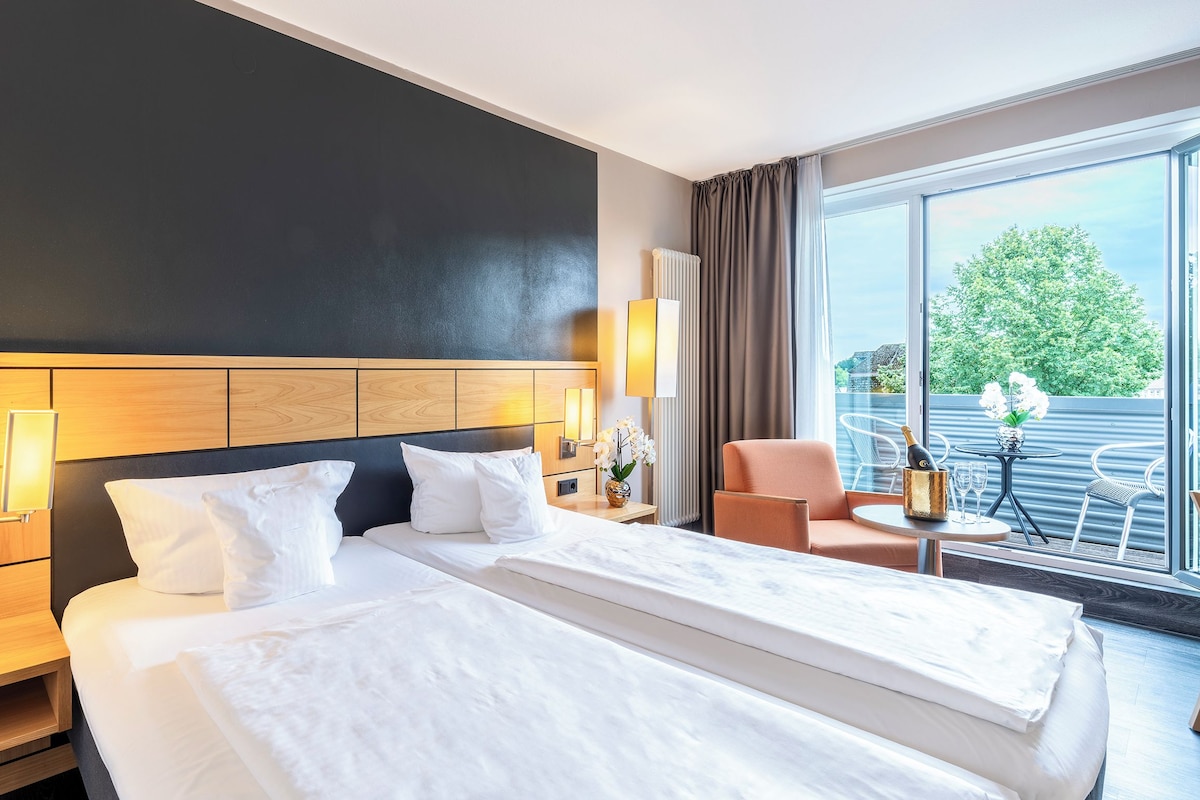 Ein Hotelzimmer mit einem großen Bett, einem Nachttisch mit Blumenvase, einem Sessel und Blick ins Freie durch große Fenster.