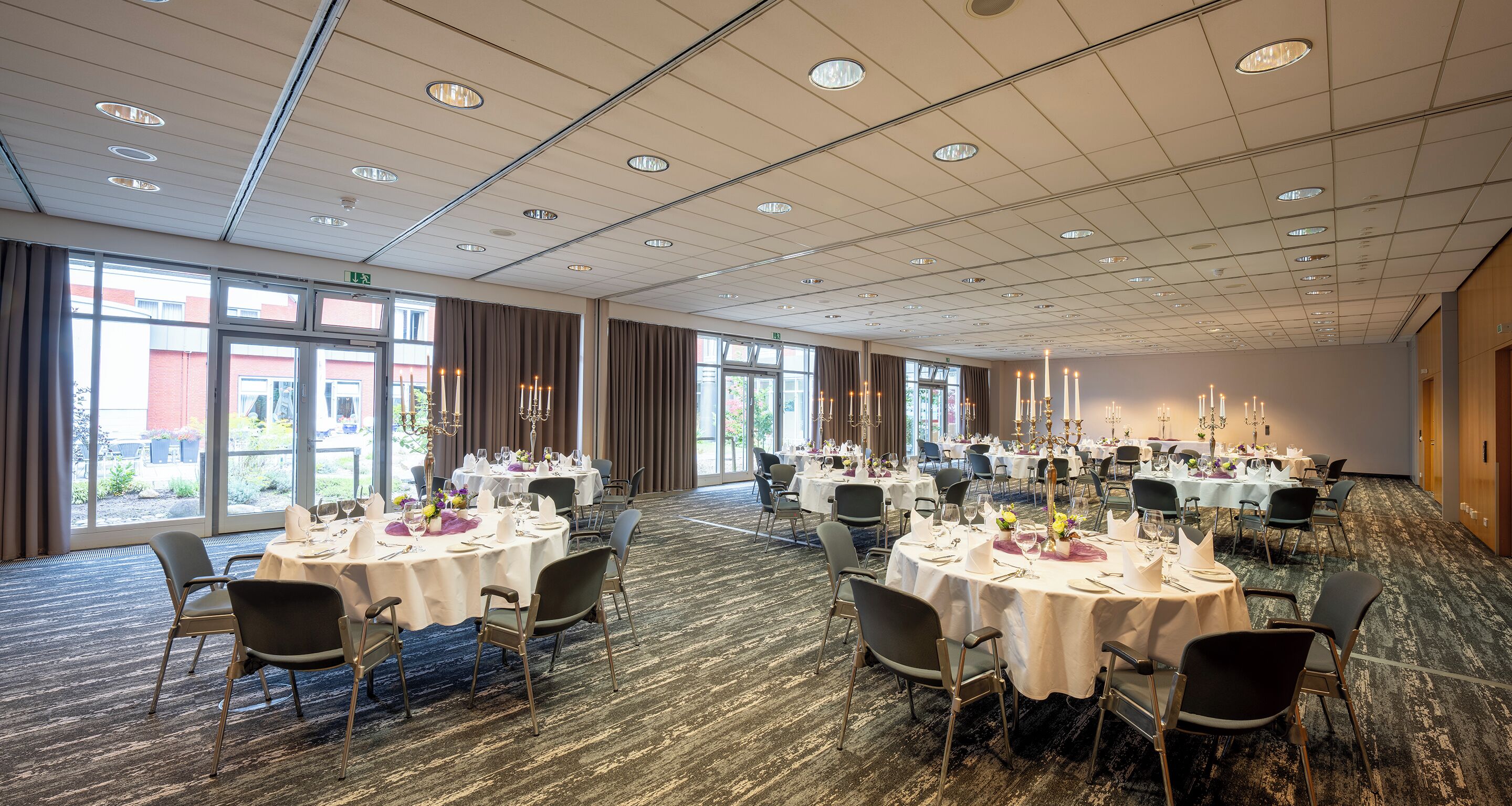 A spacious banquet hall with round tables set for dining, large windows, and a high ceiling with recessed lighting.