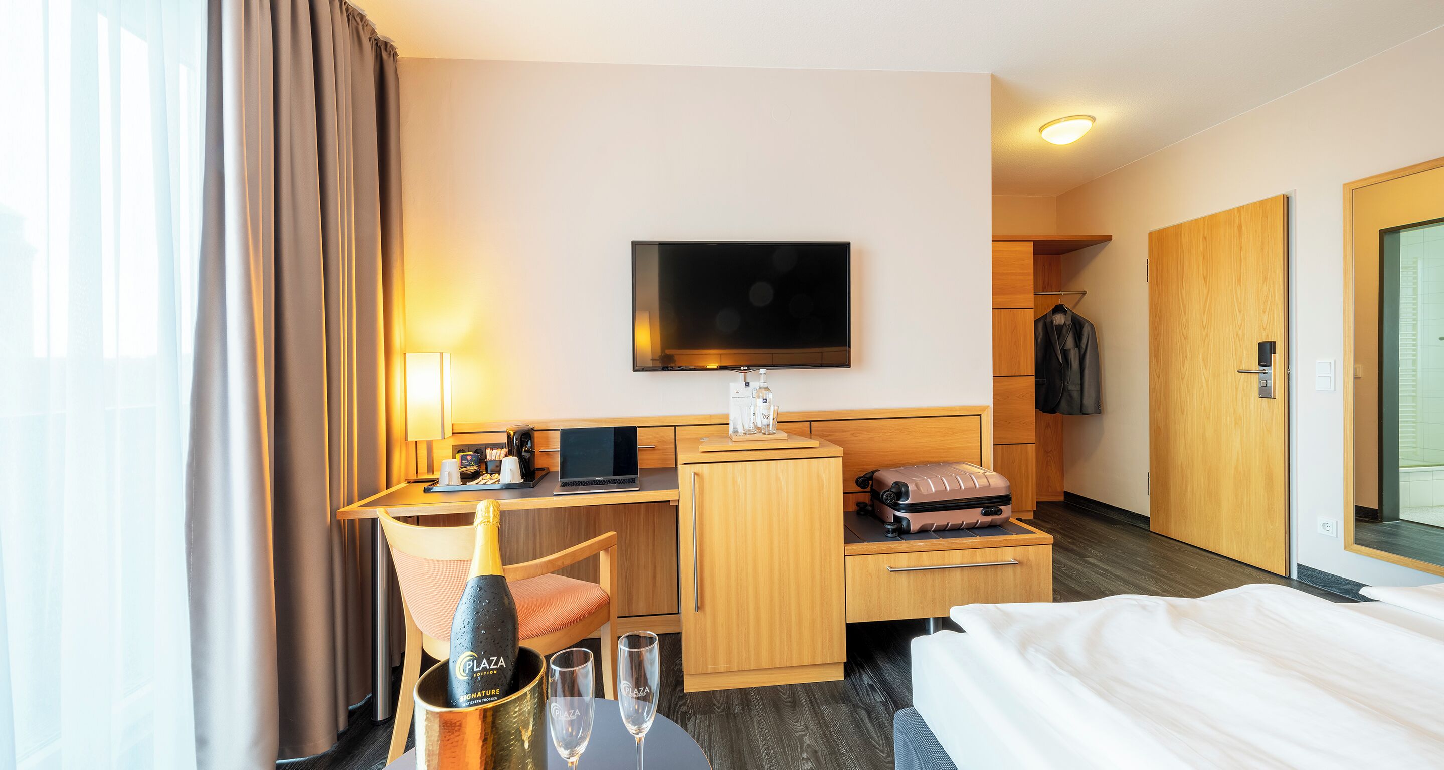 A hotel room with a bed, a desk with a laptop, a television, a minibar, and a wardrobe.