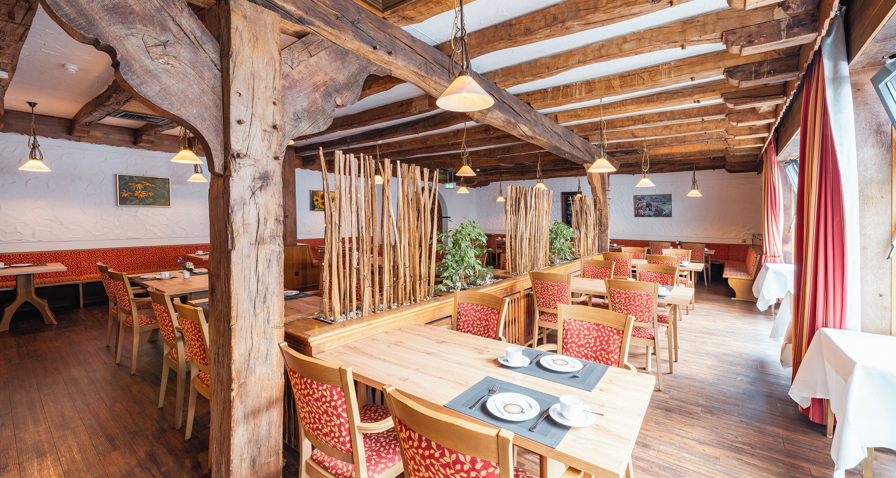 A restaurant with wooden tables and chairs, a high ceiling with exposed beams, and hanging pendant lights.