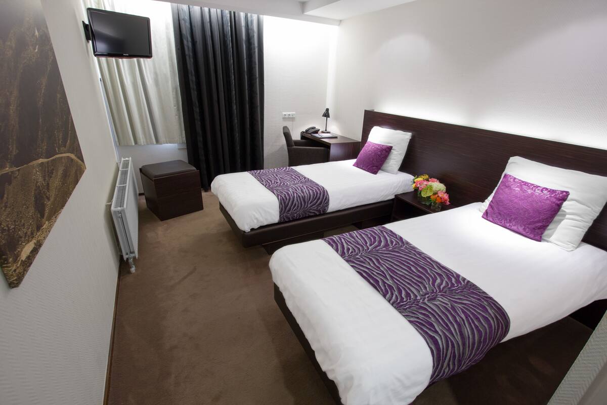 A hotel room with two beds, a desk, a chair, a TV, and a window with curtains.