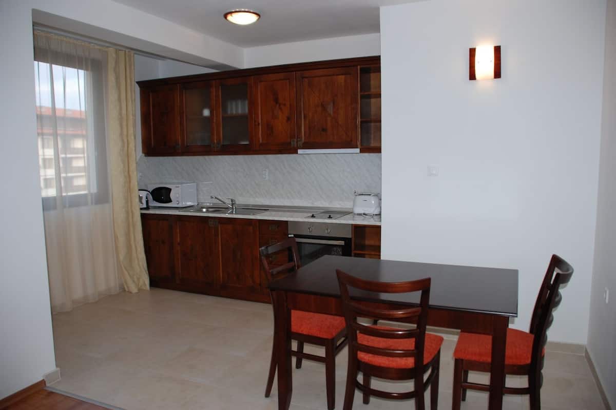 A compact kitchen with wooden cabinets, a dining table with chairs, and a window with sheer curtains.
