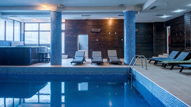 A modern indoor pool area with a clear pool, lounge chairs, and a seating area with a patterned floor. - Elegant Lodge Hotel (Bansko)