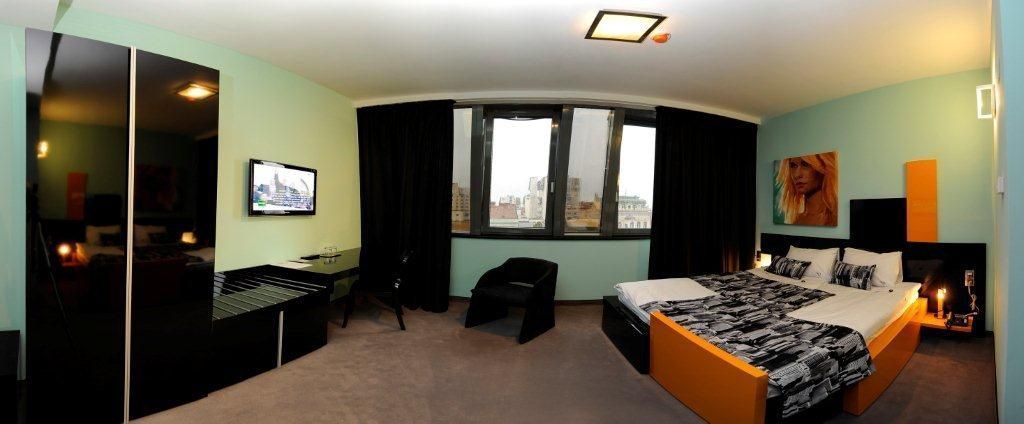 A modern hotel room with a curved bed, a television, a desk, and a view of the city through large windows.