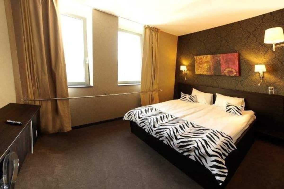 A hotel room with a large bed, a desk, a mirror, a TV, and a window with curtains.