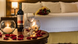 Quarto de hotel com cama, mesa de cabeceira, uma garrafa de vinho, copos, vela e pétalas de rosa.