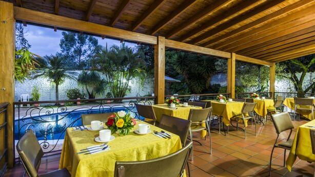 Uma área de refeições ao ar livre com toalhas de mesa amarelas, cadeiras de metal e vista para árvores e uma piscina.