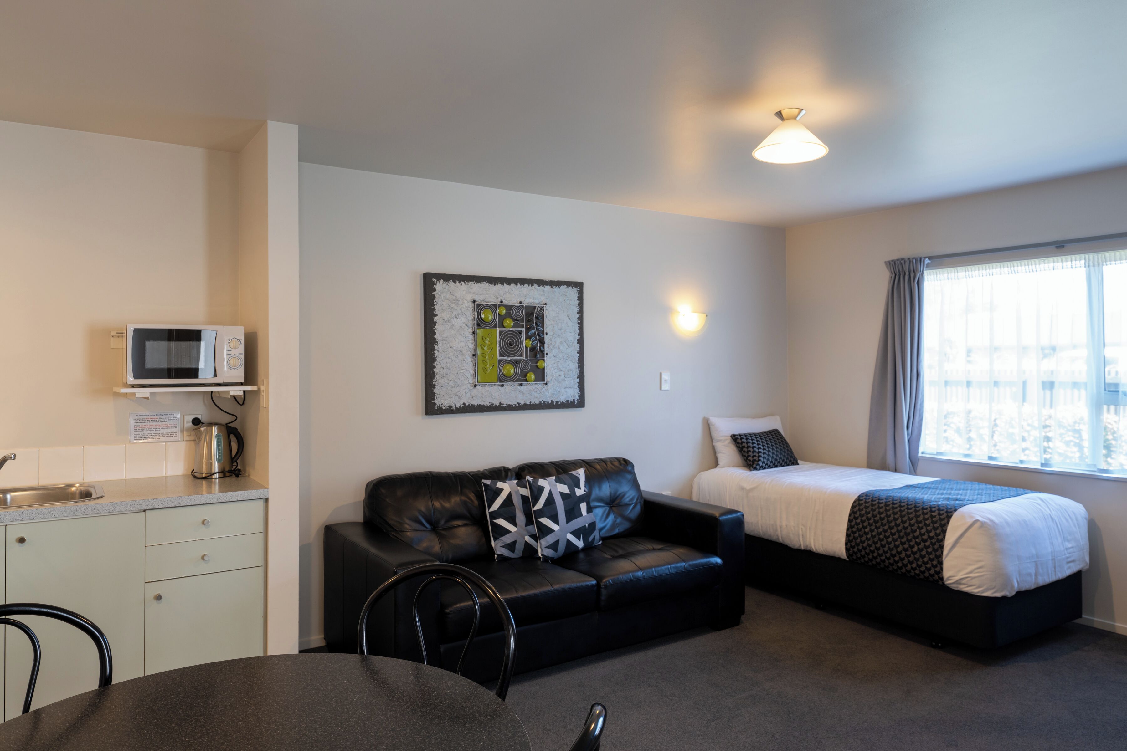 A compact room with a kitchenette, a black leather sofa, a bed, and a dining table.