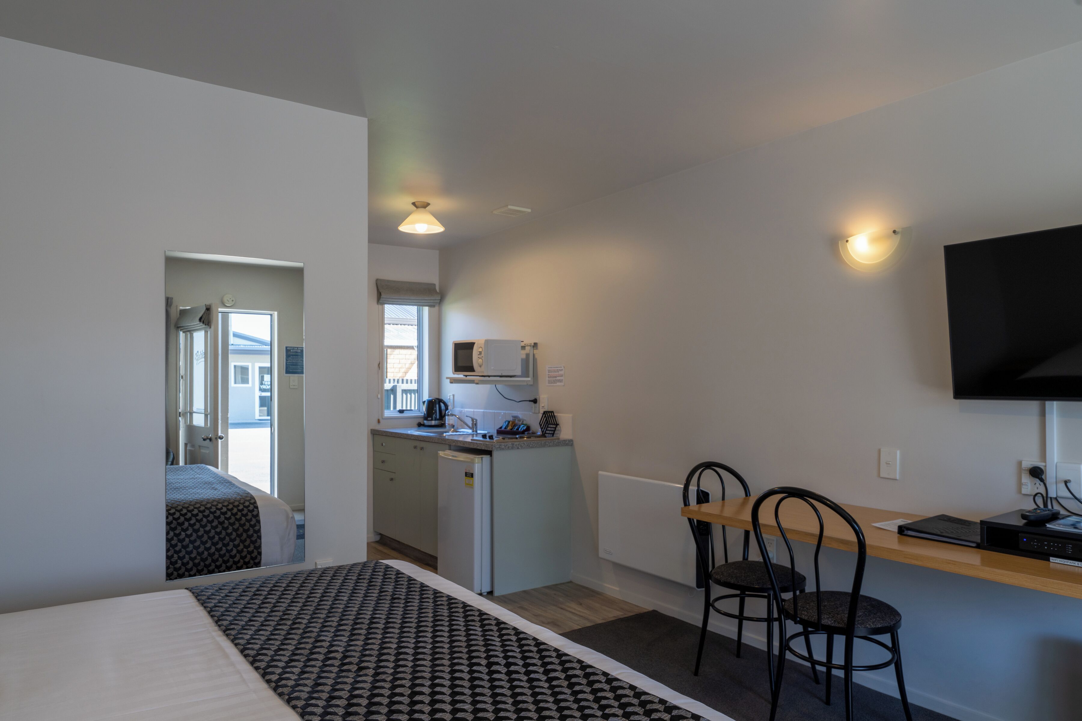 A compact hotel room with a bed, a small kitchenette, a TV, and a seating area with two chairs.